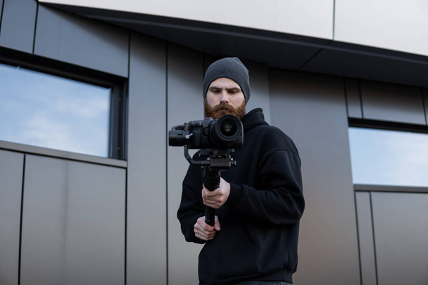 Bearded Professional videographer in black hoodie holding professional camera on 3-axis gimbal stabilizer. Filmmaker making a great video with a professional cinema camera. Cinematographer. - Φωτογραφία, εικόνα