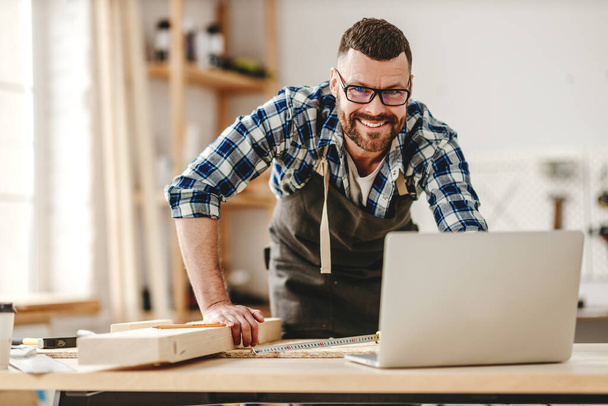 Adulto carpinteiro masculino em óculos e avental procurando informações no laptop enquanto trabalhava em worksho luz moderna
 - Foto, Imagem