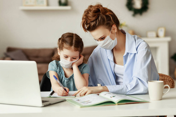 Erschöpfte Frau in medizinischer Maske sitzt am Tisch und beobachtet fleißiges Mädchen beim gemeinsamen Hausaufgabenschreiben während einer Pandemie - Foto, Bild