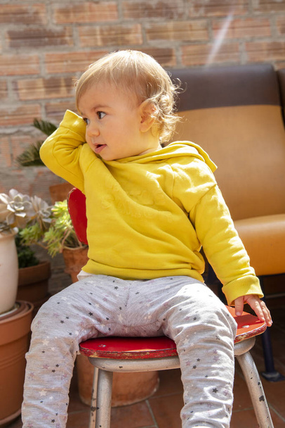 linda niña con cabello rubio y sudadera con capucha amarilla sentada en una silla roja en un patio de jardín
 - Foto, Imagen