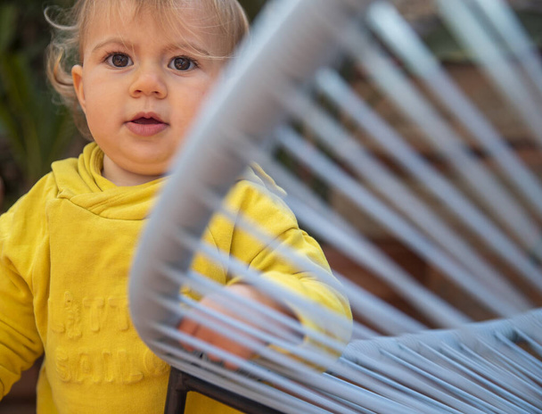 linda niña con cabello rubio y sudadera con capucha amarilla jugando en un patio de jardín disparado a través de una silla de cuerda
  - Foto, Imagen