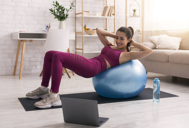 Stay home. Young girl doing exercises with fitness ball while watching online tutorials on laptop - Foto, Imagen