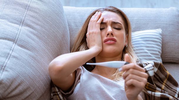 Woman has headache. She lying on sofa and holds thermometer - Photo, image