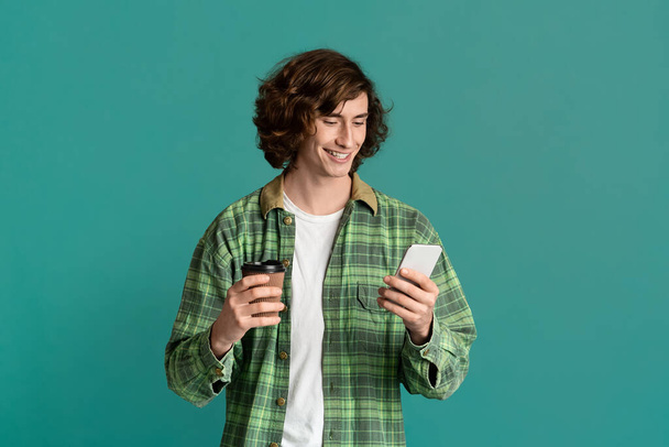 Szczęśliwy młody facet ze smartfonem i filiżanką kawy komunikujących się online przez telefon komórkowy, turkusowe tło - Zdjęcie, obraz