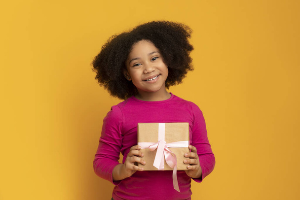 Smiling little black girl holding gift box, posing over yellow background - Foto, afbeelding