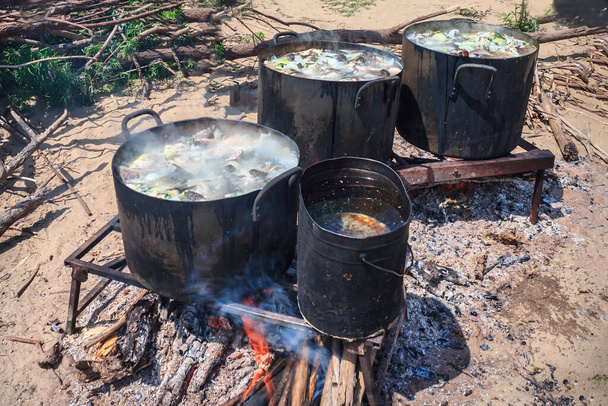 Cooking lunch in a hike, a fire on the sandy shore. Large pans with fish soup. Firewood, embers, bright fire. Hot sunny day, summer. - Photo, Image