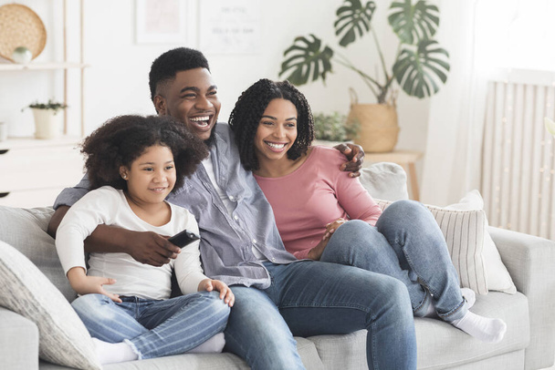 Черная семья из трех человек отдыхает на диване, вместе смотрит телевизор
 - Фото, изображение