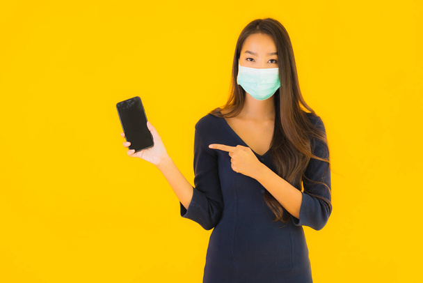 Портрет красивая молодая азиатская женщина с маской и смартфона или мобильного телефона на желтом изолированном фоне
 - Фото, изображение