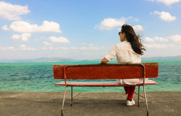 Азиатка носит белую рубашку, красные длинные брюки и сидит на красной скамейке у моря
 - Фото, изображение