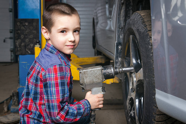 Młody chłopak, młody pracownik samochodowy, zmienia oponę za pomocą klucza pneumatycznego w garażu stacji paliw. Dziecko uczy się mechaniki zmieniającej zawód w serwisie naprawy samochodów. - Zdjęcie, obraz