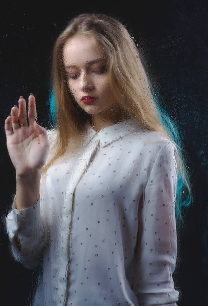 A beautiful girl in a white shirt is sad behind a glass with water drops, a photo of a glamorous young, slender woman behind a blind glass - 写真・画像