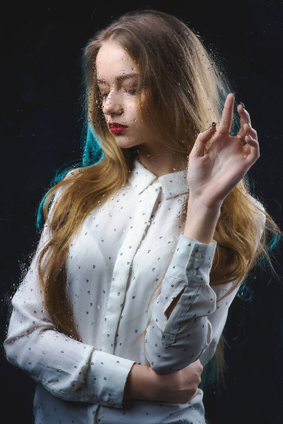 A beautiful girl in a white shirt is sad behind a glass with water drops, a photo of a glamorous young, slender woman behind a blind glass - Valokuva, kuva
