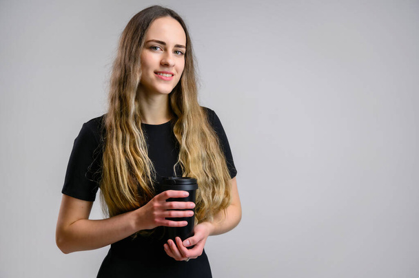 Portrait of a pretty young woman in a black dress with long hair. A model stands on a white background with a cup of coffee in her hands. - Photo, Image