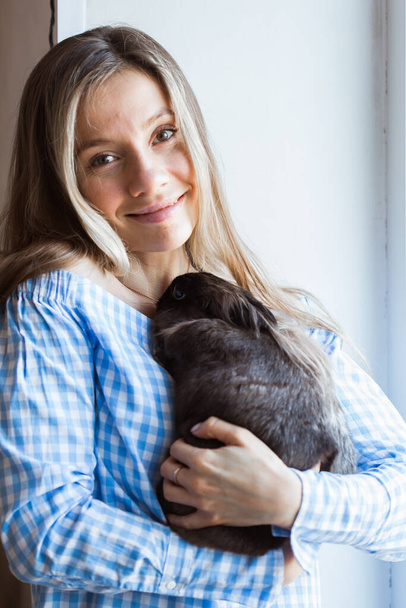 Mascotas y concepto de Pascua - Chica atractiva abrazando conejo marrón en casa
. - Foto, Imagen