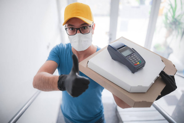 Deliveryman with protective medical mask holding pizza box and POS wireless terminal for card paying - days of viruses and pandemic, food delivery to your home. - Фото, изображение