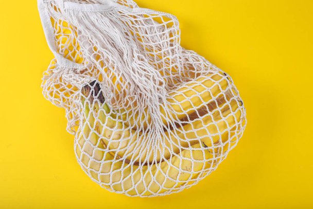 黄色の背景に新鮮なバナナと再利用可能なショッピングバッグ。生態学的概念。メッシュショッピングコットンバッグのトップビュー。環境への配慮とプラスチックの除去. - 写真・画像