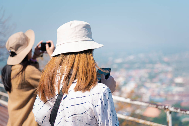 アジアのかわいい女性の帽子はリラックスし、山の上の海辺の都市景観の視点でスマートフォンを使用し、コンセプト旅行、休暇、生活の中でのレジャーで幸せと自由の感情 - 写真・画像