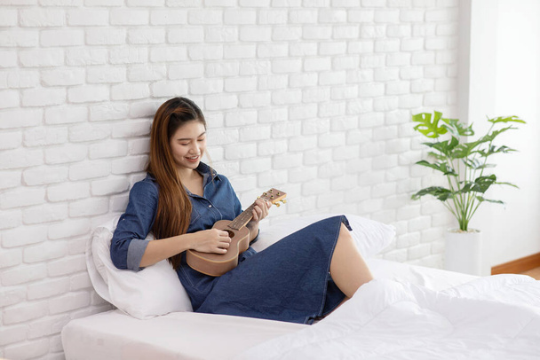 Attractive Asian young woman playing Ukulele on bed in cozy bedroom and sing a song relaxing with music,Recreation in holidays Concept - Фото, зображення