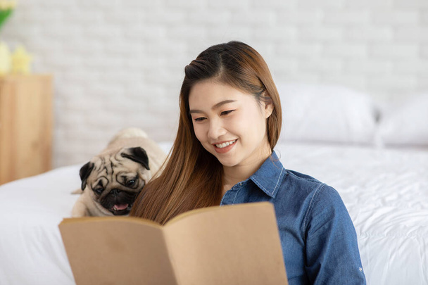 Attractive Asian young woman reading book while her dog pug breed in cozy bedroom relaxing and comfortable in holidays,Recreation with dog Concept - Foto, Imagem