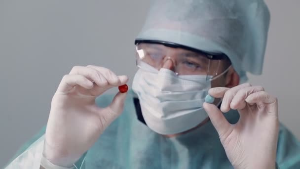 Doctor dressed in a surgical gown in a medical mask with glasses points at two tablets of red and blue. Doctor holding two pills in white medical gloves. - Filmagem, Vídeo