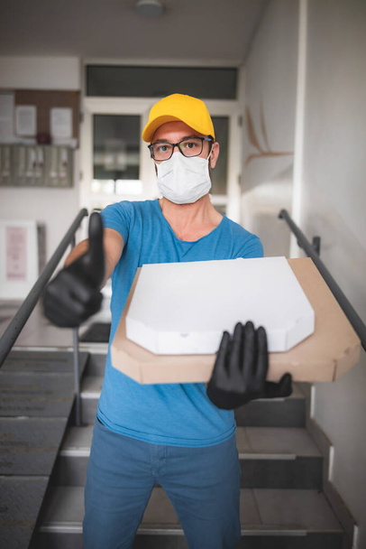 Lieferant mit medizinischer Schutzmaske hält Pizzaschachtel - Tage der Viren und Pandemie, Essenslieferung nach Hause. - Foto, Bild