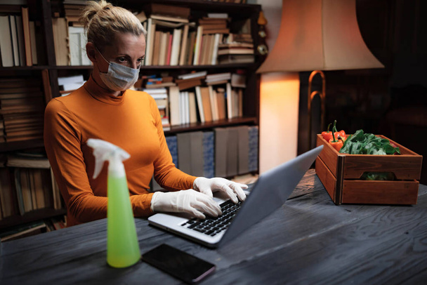 Vrouw met beschermend masker tegen virussen en ziekteverwekkers die thuis laptop gebruikt, levensmiddelen, levensmiddelen en benodigdheden bestelt via internet. - Foto, afbeelding