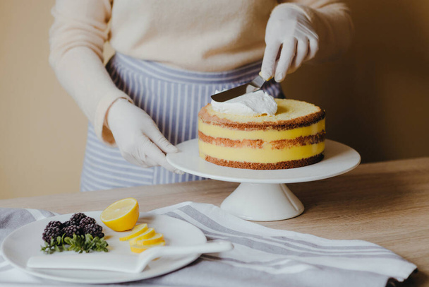 Νοικοκυρά στην κουζίνα μαγειρεύοντας ένα κέικ λεμονιού με φρούτα, βατόμουρο, λεμόνι και μέντα σε κίτρινο φόντο κατά τη διάρκεια της καραντίνας. Συνταγή της βιολογικής σπιτικό κέικ λεμονιού. Μαγειρική στο σπίτι. - Φωτογραφία, εικόνα