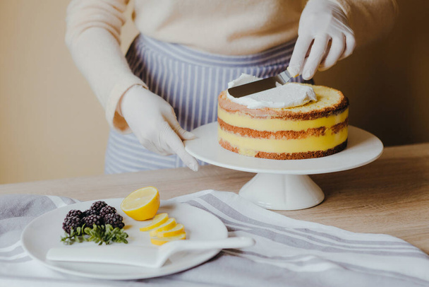 Housewife in the kitchen cooking a lemon cake with fruits, blackberry, lemon and mint on yellow background during quarantine. Recipe of organic homemade lemon cake. Cooking at home. - Photo, image