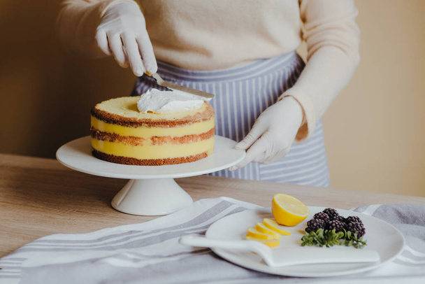 Νοικοκυρά στην κουζίνα μαγειρεύοντας ένα κέικ λεμονιού με φρούτα, βατόμουρο, λεμόνι και μέντα σε κίτρινο φόντο κατά τη διάρκεια της καραντίνας. Συνταγή της βιολογικής σπιτικό κέικ λεμονιού. Μαγειρική στο σπίτι. - Φωτογραφία, εικόνα