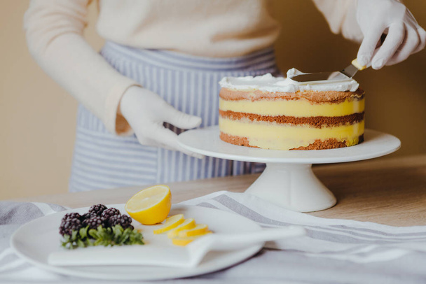 Housewife in the kitchen cooking a lemon cake with fruits, blackberry, lemon and mint on yellow background during quarantine. Recipe of organic homemade lemon cake. Cooking at home. - 写真・画像