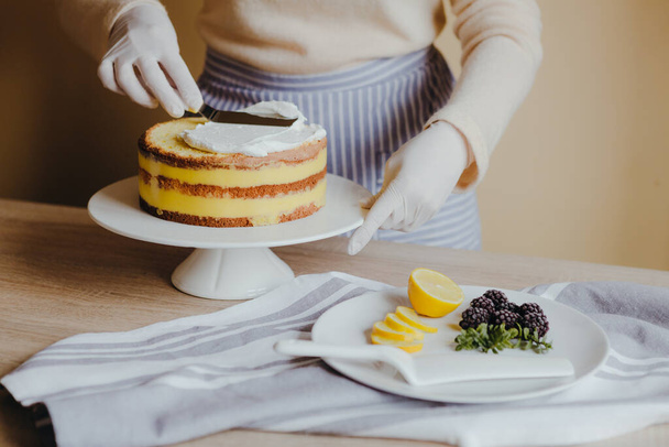 Huisvrouw in de keuken kookt een citroentaart met fruit, bramen, citroen en munt op gele achtergrond tijdens quarantaine. Recept van biologische zelfgemaakte citroentaart. Koken thuis. - Foto, afbeelding