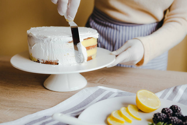 Hausfrau breitet das Sahnehäubchen aus, um die Spitze des Kuchens zu bedecken. Hausfrau glättet Oberfläche mit Spachtel, Nahaufnahme ausgeschnittenes Foto. Freizeit in Quarantäne. - Foto, Bild