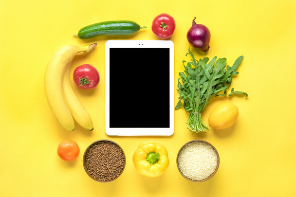 różne rodzaje zdrowej żywności - gryka, ryż, żółta papryka, pomidory, banany, sałata, zieleń, ogórek, cebula, tabletka z czarnym ekranem na żółtym tle Top view Flat lay Online shopping Mockup - Zdjęcie, obraz