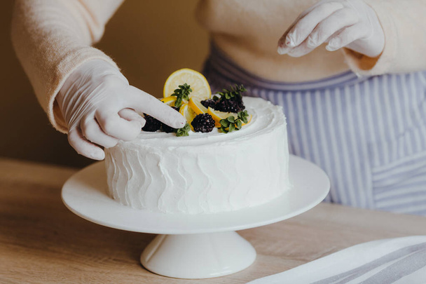 Housewife in the kitchen decorates a lemon cake with fruits, blackberry, lemon, mint during quarantine. Recipe of organic homemade lemon cake - Photo, Image