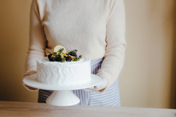 Femme au foyer dans la cuisine tenant un gâteau au citron avec des fruits, mûres, citron, menthe pendant la quarantaine. Recette de gâteau au citron maison. Concept de cuisine à la maison
. - Photo, image