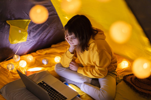 Young girl student using laptop for homeschooling in a home made tent with glowing light balls. - Фото, изображение