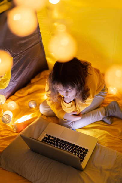 Юная студентка использует ноутбук для домашнего обучения в домашней палатке с светящимися шарами
. - Фото, изображение