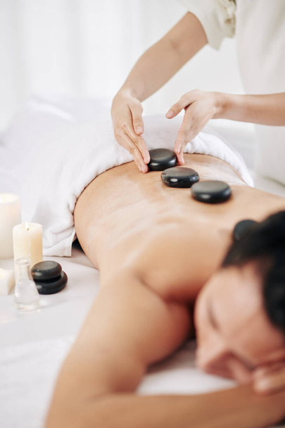 Masaż terapeuta wprowadzenie ciepłe kamienie masażu wzdłuż kręgosłupa klienta salon spa - Zdjęcie, obraz