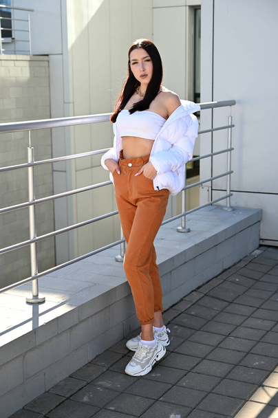 Vertical Full length photo outdoors of a young brunette girl in beige trousers and a white jacket on the background of the railing of the building s stairs in the city. Made on a sunny spring day. - Фото, изображение