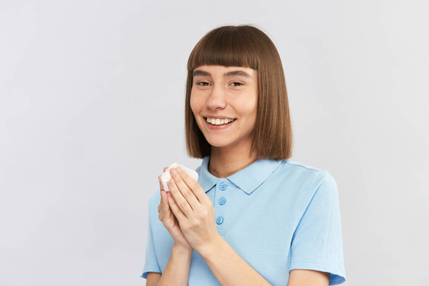 χαρούμενη νεαρή κοπέλα κρατώντας λευκό ιστό μετά φυσώντας μύτη και κοιτάζοντας κάμερα με χαμόγελο - Φωτογραφία, εικόνα