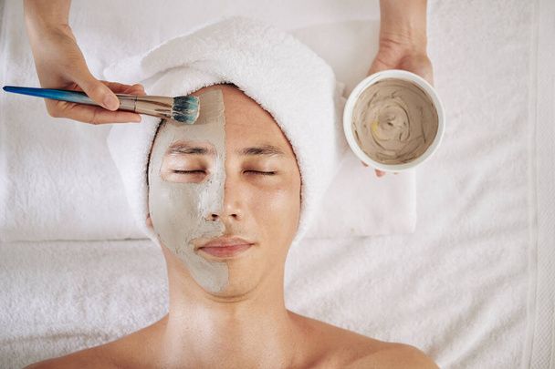 Kosmetolog nanáší masku z dřevěného uhlí na polovinu obličeje dospělého muže, pohled shora - Fotografie, Obrázek