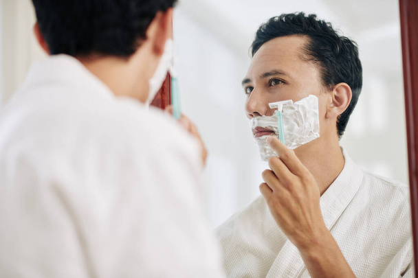 Hombre serio de raza mixta mirando el espejo del baño y afeitándose la barba
 - Foto, imagen