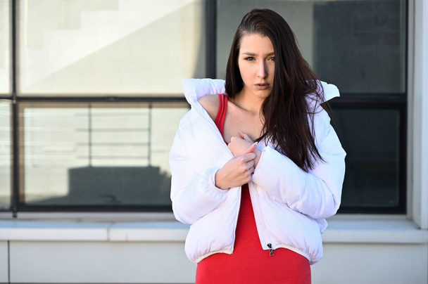 Outdoors portrait of a fashionable young brunette girl in a white jacket stands against the background of a building wall. - Фото, изображение