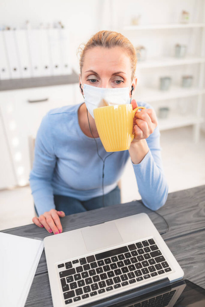 Online leraar met een beschermend masker bijles over het internet. Problemen met het drinken van koffie in de ochtend. - Foto, afbeelding