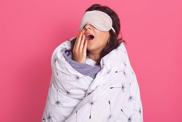 Close up portrait of brunette woman wearing blindfold and wrapped in white blanket, standing and yawning with widely opened mouth, poses isolated over pink background. Stay home during quarantine. - Photo, image