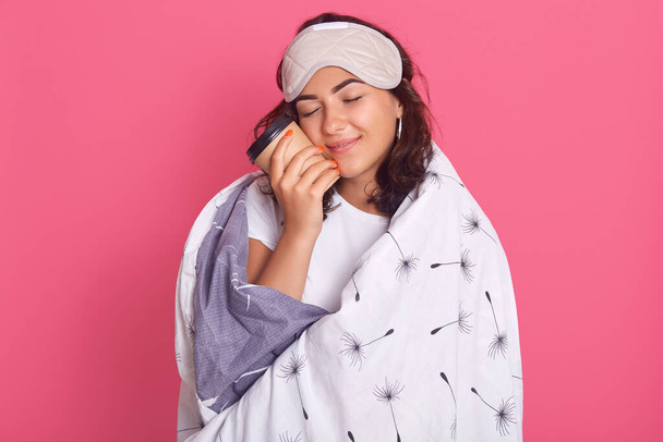 Close up portrait of dark haired woman posing with closed eyes, holding takeaway hot coffee, enjoying stay home during quarantine, wrapped in white blanket, isolated over pink stdio background. - Фото, изображение