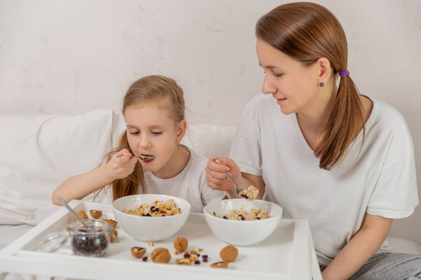 Happy mom and daughter have healthy breakfast on bed in a light bedroom on a sunny morning. Oatmeal porridge with nuts and raisins. Healthy food and family relationships concept. Good mood. - Photo, image
