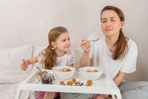 Happy mom and daughter have healthy breakfast on bed in a light bedroom on a sunny morning. They have fun and enjoy. Healthy food and family relationships concept. Good mood. Space for text. - Photo, image