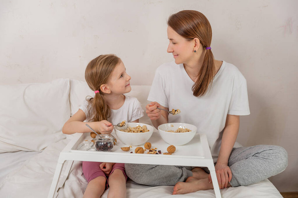 幸せな母親と娘は晴れた朝にベッドで健康的な朝食を持っています。彼らはお互いを見て笑顔。健康食品の概念。気分がいい。テキストのためのフリースペース. - 写真・画像