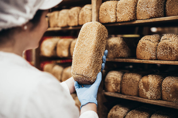 Jonge vrouwelijke werkneemster die in de bakkerij werkt en brood op de plank zet - Foto, afbeelding